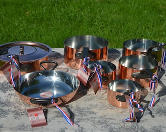 May include: A collection of copper cookware with stainless steel interiors. The set includes pots, pans, and a lidded saucepan, all displayed on a patterned tablecloth. Each piece has a black handle and is adorned with a red, white, and blue ribbon.