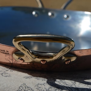 Puede incluir: Una sartén redonda de cobre con un interior plateado pulido y dos asas de latón fijadas con remaches de cobre. El lado exterior de la sartén presenta el texto estampado MADE IN FRANCE. La sartén tiene una profundidad poco profunda y lisa.