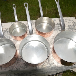 Può includere: Una collezione di cinque casseruole con fondo in rame e lunghi manici argentati. Le pentole sono disposte su una tovaglia fantasia, mettendo in risalto i loro interni lucidi e gli esterni in rame a contrasto. I manici hanno un anello per appendere.