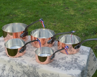 May include: Set of five copper saucepans with black handles and a shiny finish. Each saucepan has a blue, white, and red ribbon tied to the handle.
