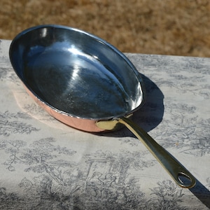 May include: An oval-shaped copper skillet featuring a polished silver-toned interior lining and a long, smooth brass handle. The pan has a warm metallic exterior finish and a sturdy, riveted connection where the handle meets the main body.