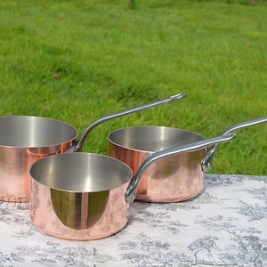 Pode incluir: Três panelas de cobre com alças de prata. As panelas estão aninhadas em uma toalha de mesa branca com um padrão floral.