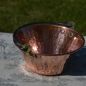May include: A small, copper-colored bucket with a hammered texture. It has a black handle and brass-colored hardware. The bucket is sitting on a patterned surface, with a green background.