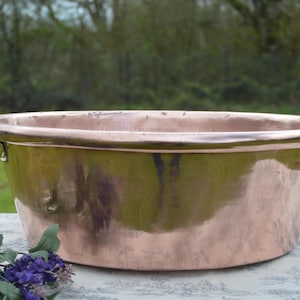 Pode incluir: Uma bacia de cobre com duas alças de latão. A bacia tem uma forma oval e um acabamento polido, refletindo o ambiente. As alças são curvas e presas às laterais. A bacia é provavelmente usada para fins decorativos ou funcionais.