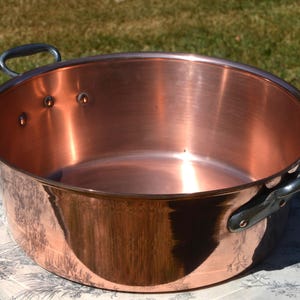 Op de afbeelding: Een ronde, gepolijste koperen kookpan met een wijde opening en twee donkere metalen handgrepen die met zichtbare klinknagels aan de zijkanten zijn bevestigd. De binnen- en buitenoppervlakken zijn glad en reflecterend met een warme, oranjebruine kleur.