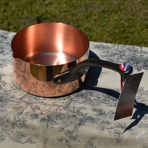 May include: A copper saucepan with a black handle. The saucepan is sitting on a white tablecloth with a floral pattern.