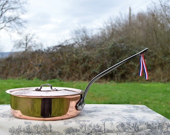 May include: A large copper pot with a lid and a long, curved handle. The pot is polished and shiny, and the handle is made of black metal. The pot is sitting on a table with a patterned tablecloth.