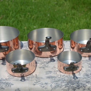 May include: A set of five round copper saucepans with silver-toned interiors and dark, curved metal handles. The pans are arranged in a group, featuring polished copper exteriors and sturdy handles attached with two visible rivets on each side.