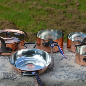 May include: Set of five copper pots with black handles and lids. The pots are arranged on a white tablecloth with a floral pattern. The pots are all different sizes and have a shiny, polished finish.