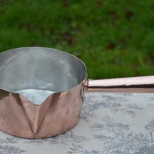 Puede incluir: Una cacerola de cobre con un mango largo. La cacerola tiene un interior brillante y pulido y un exterior ligeramente abollonado.