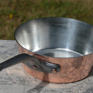 May include: A copper-clad saucepan with a silver interior and a dark handle. The pan is set on a patterned surface, with a blurred green background.