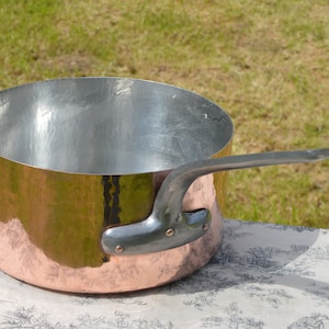 Puede incluir: Una olla de cobre con un acabado martillado y un asa de plata. La olla está vacía y se encuentra sobre un mantel blanco con un patrón floral.