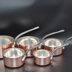 May include: Set of four copper saucepans with silver handles. The saucepans are nested together in order of size, with the largest saucepan at the bottom and the smallest saucepan at the top.