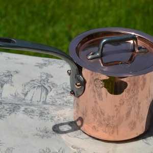 Op de afbeelding: Een koperen steelpan met een deksel en een lange handgreep. De pot heeft een glanzend, reflecterend oppervlak en een donkere metalen handgreep. Het deksel heeft ook een donkere metalen handgreep.