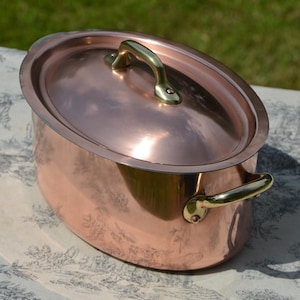 May include: An oval copper pot with a polished finish, featuring a matching lid and two brass side handles. The lid is topped with a single brass handle, and the entire piece exhibits a reflective surface that captures light across its curved body.