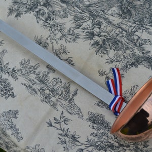 May include: A copper ladle with a long, silver handle. The handle has a blue, white, and red ribbon tied to it.