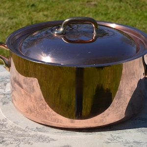 Pode incluir: Uma panela de cobre com tampa e duas alças laterais. A panela tem uma superfície brilhante e reflexiva e uma alça de latão na tampa. A panela está sobre uma toalha de mesa estampada.