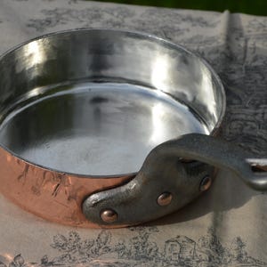 May include: A copper saucepan with a black iron handle. The saucepan has a hammered finish and is shiny and reflective.