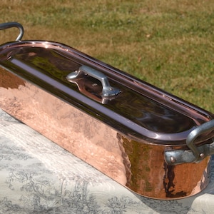Pode incluir: Uma panela de peixe de cobre, de formato oval e alongado, com acabamento polido. O recipiente possui duas alças laterais de metal rebitadas e uma tampa correspondente com uma alça central de metal, refletindo a luz na sua superfície.