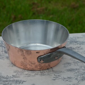 Op de afbeelding: Een koperen steelpan met een zilveren binnenkant en een grijze handgreep. De pan staat op een patroonkleed, met een groene achtergrond. Gemaakt in Frankrijk.