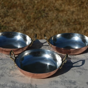 May include: Three round serving dishes with polished copper exteriors and shiny silver-toned interiors. Each dish features two small, curved brass-colored metal handles attached to the sides, providing a contrast to the warm copper finish.