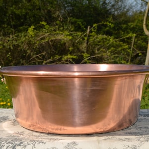 May include: A large, round, polished copper basin featuring a wide, flat rim and two sturdy, curved brass handles attached to opposite sides. The exterior surface of the metal vessel is smooth and reflective, showcasing a warm, metallic orange-brown hue.