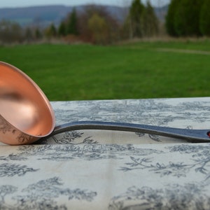 May include: A copper ladle with a long, black, hammered handle. The ladle is resting on a patterned tablecloth with a blue, white and red ribbon tied to the handle.