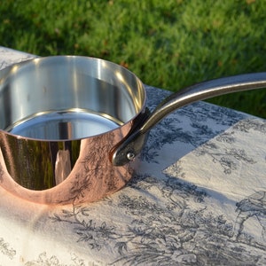 Peut inclure: Une casserole en cuivre avec un intérieur argenté et une poignée en métal foncé. La casserole est posée sur une nappe à motifs. La poignée est rivetée sur le côté de la casserole. Le diamètre de la casserole est d'environ 15 cm.