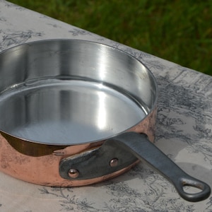 May include: A copper-bottomed saucepan with a stainless steel interior and a riveted metal handle. The pan is round and sits on a patterned surface, with a green background.