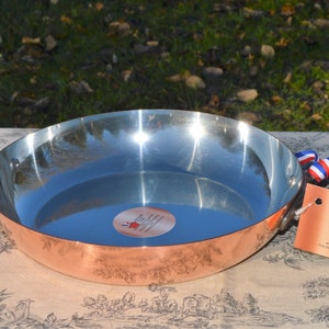 May include: A copper pan with a shiny silver interior and two black handles. The pan is sitting on a patterned tablecloth. A tag with the text "Normandy Kitchen Copper" is attached to the handle.