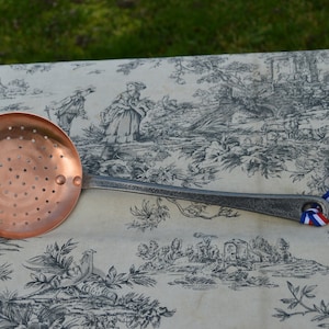 May include: A copper slotted spoon with a silver handle and a blue, white, and red ribbon tied around the handle.