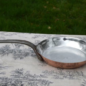 以下が含まれることがあります： 長いダークメタルハンドルが付いた銅底のフライパン。フライパンの内側は銀色で、外側は温かみのある銅色です。フライパンは、緑色の背景に、花柄のデザインの模様の生地の上に置かれています。