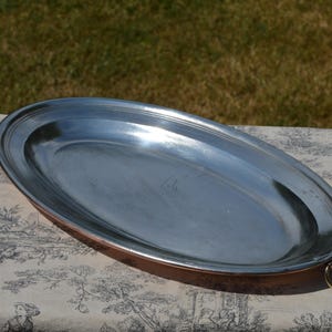 May include: An oval-shaped serving platter featuring a polished silver-toned interior surface and a copper-toned exterior rim. The platter includes a small, circular metal ring handle attached to one side, providing a functional grip for handling.