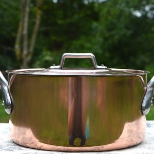 Peut inclure: Une casserole de cuisine de couleur cuivre avec un couvercle et deux poignées incurvées. La casserole a une surface brillante et réfléchissante et une poignée argentée sur le couvercle. La casserole est posée sur une surface blanche avec un fond vert.