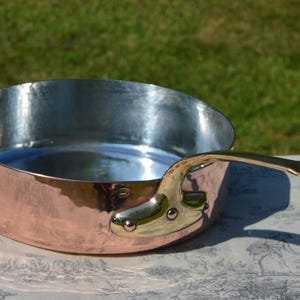 May include: A copper saucepan with a silver interior and a brass handle. The pan is round with a wide base and a curved handle attached with rivets. The background is a blurred green lawn.