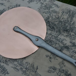 Può includere: Un coperchio per pentola in rame con un manico in argento. Il coperchio è rotondo e ha una superficie liscia. Il manico è lungo e ha un anello all'estremità.