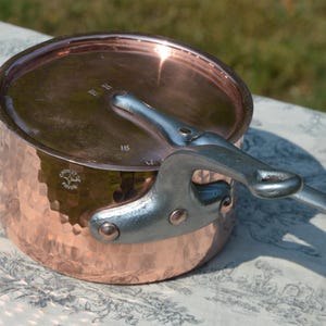 May include: A copper cooking pot with a hammered finish and a lid. The pot has a silver-coloured handle. The pot is sitting on a patterned surface. The pot is likely used for cooking.