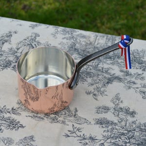 May include: A copper saucepan with a silver interior and a long, dark handle. The pan has a decorative design on the exterior and is adorned with red, white, and blue ribbons tied to the handle. The pan is sitting on a patterned tablecloth.