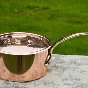 Peut inclure: Une casserole en cuivre avec un couvercle et une poignée en laiton. La casserole est brillante et reflète la lumière. La casserole est posée sur une nappe blanche à motifs floraux.