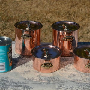 May include: A set of four polished copper pots with lids and brass handles, arranged alongside a single blue and silver metal food can. The pots vary in height and diameter, featuring metallic surfaces that reflect light and surrounding colours.