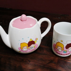 Puede incluir: Una tetera de cerámica blanca con tapa rosa y una taza a juego. Ambas presentan una ilustración de dibujos animados de dos personajes con cabello amarillo y marrón, rodeados de estrellas y el texto "Little Twin Stars".