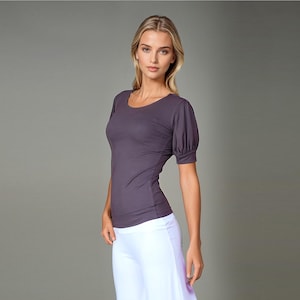 May include: A dark purple scoop-neck top with short, puffed sleeves. The fitted top is paired with white pants. The model is standing in front of a gray background.
