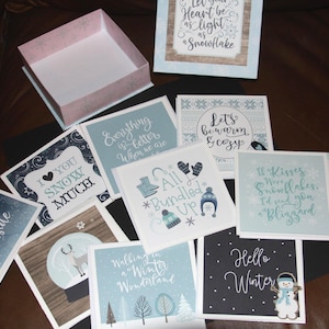 May include: A collection of winter-themed greeting cards with various designs and quotes, including "Baby it's Cold Outside" and "Hello Winter." The cards are in shades of blue, white, and brown, with a decorative box in the background.
