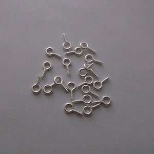 May include: A collection of small, silver-colored screw eye pins scattered on a gray surface. Each pin has a looped eye and a threaded screw end, designed for attaching to various materials. These are commonly used in jewelry making and crafts.