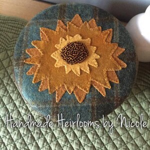 May include: A handmade felt sunflower with a brown bead center, on a blue and green plaid background. The text "Handmade Heirlooms by Nicole" is visible below the sunflower.