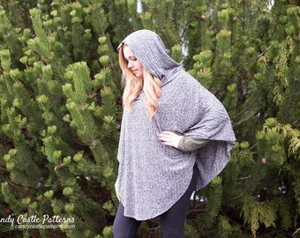 May include: A grey hooded poncho with a ribbed texture. The poncho is being worn by a person with long blonde hair. The person is standing in front of a green bush.