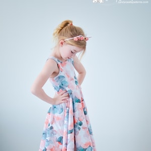 Op de afbeelding: Een jong meisje draagt een mouwloze jurk met een aquarelprint van roze, blauwe en groene cirkels. Ze staat op een houten vloer en heeft een roze bloemenhoofdband op.