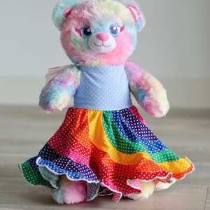 May include: A rainbow tie-dye teddy bear wearing a blue and white polka dot dress with a rainbow and white polka dot skirt. The skirt has a ruffled hem.