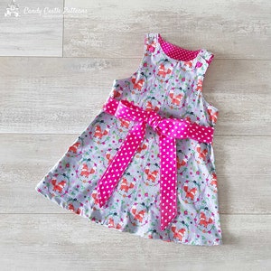 Op de afbeelding: Een grijs jurkje zonder mouwen met een roze gebloemde lintgordel. Het jurkje heeft een vos- en bloemenprint. Het jurkje is gemaakt door Candy Castle Patterns.