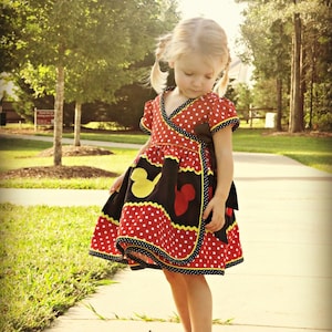 Op de afbeelding: Een rood-zwarte polkadotjurk met gele en zwarte afwerking. De jurk heeft een wikkelontwerp en is versierd met op Mickey Mouse geïnspireerde vormen. Het kind draagt zwarte schoenen. De jurk is voor een kind.